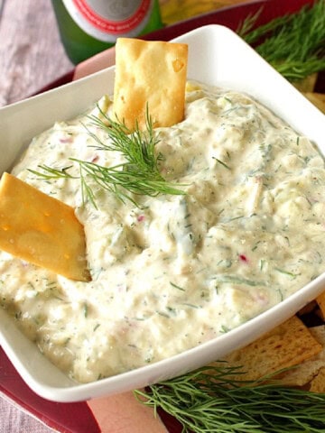 A square white bowl filled with cucumber dill dip with two crackers sticking in the dip with fresh dill sprigs on top.