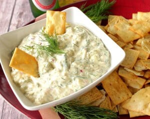A square white bowl filled with cucumber dill dip with two crackers sticking in the dip.