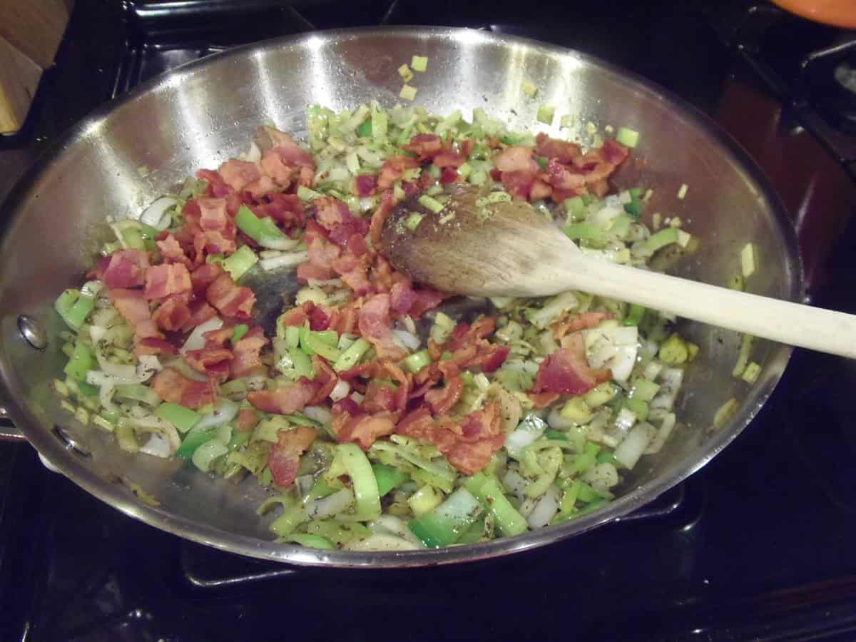 Slices of leeks and bits of bacon cooking together in a large skillet.
