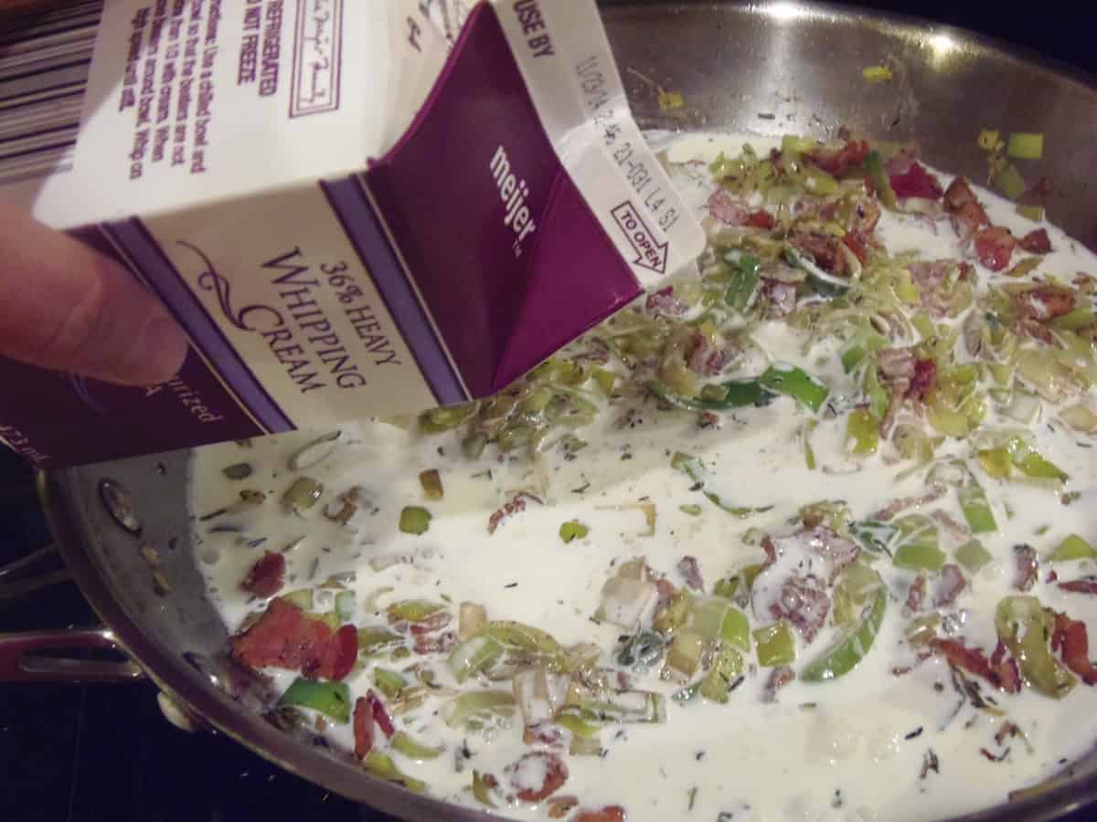 Cream being added to a skillet with bacon and leeks.