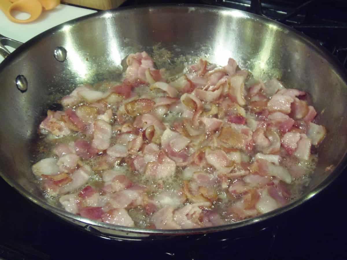 Cut up bacon frying in a large silver skillet.