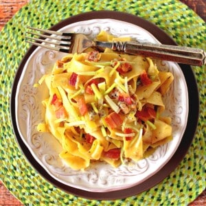 A round green placemat topped with two plates holding one serving of delicious looking pappardelle pasta with bacon and leeks.