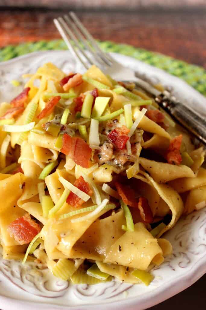 Slices of cooked leek and bacon on top of a plate of pappardelle pasta.