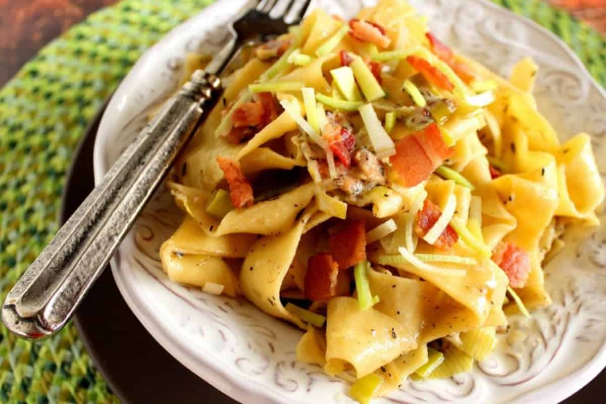 A serving of Pappardelle pasta in a bowl topped with bacon and leeks.