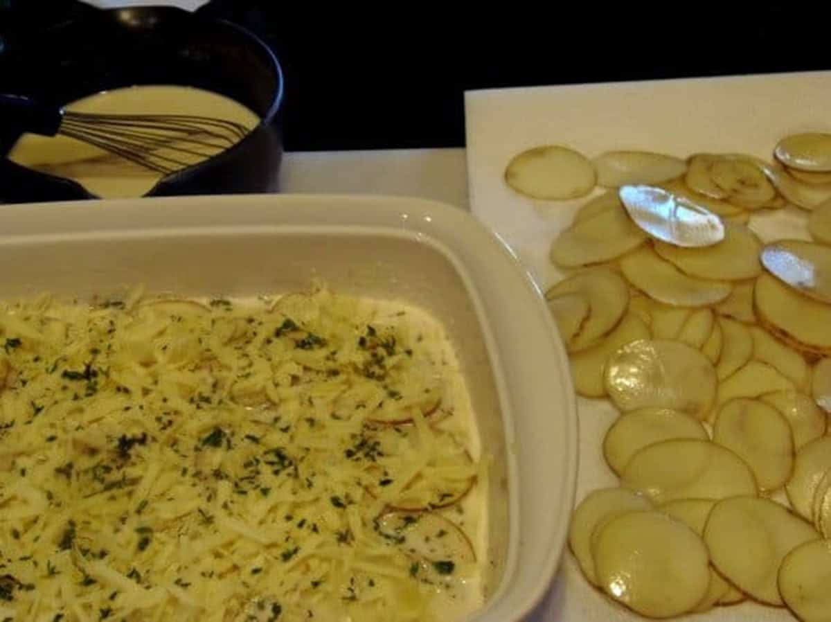 Sliced potatoes on a cutting board next to a casserole dish layering potatoes to make au gratin potatoes.