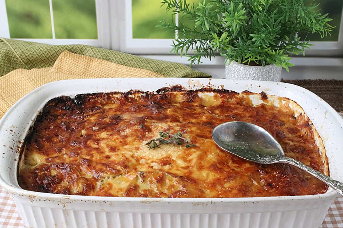 A white rectangle casserole dish filled with cheesy brown au gratin potatoes and topped with fresh thyme and a serving spoon.