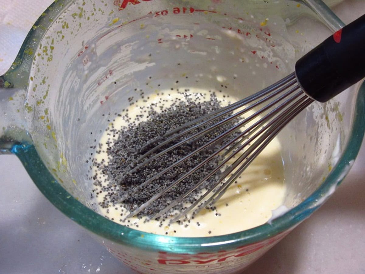 A whisk in a glass measuring cup along with mayonnaise and poppy seeds.