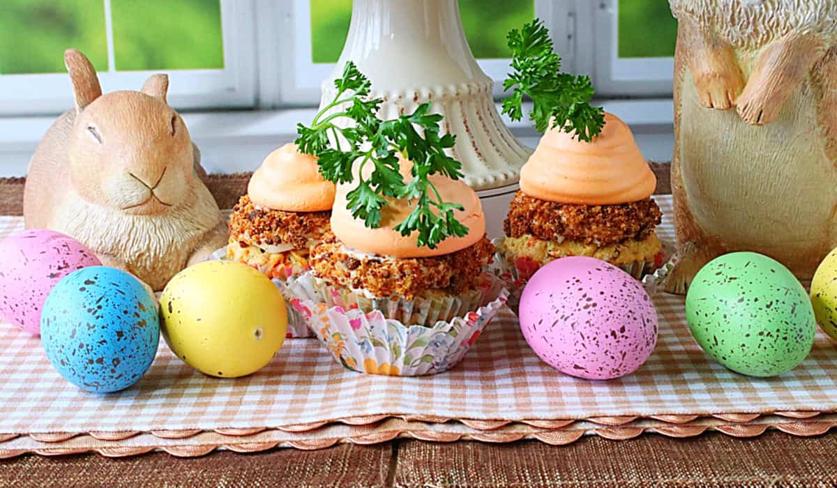 Three parsley topped meringue carrot cupcakes on a gingham checked tablecloth with Easter eggs.