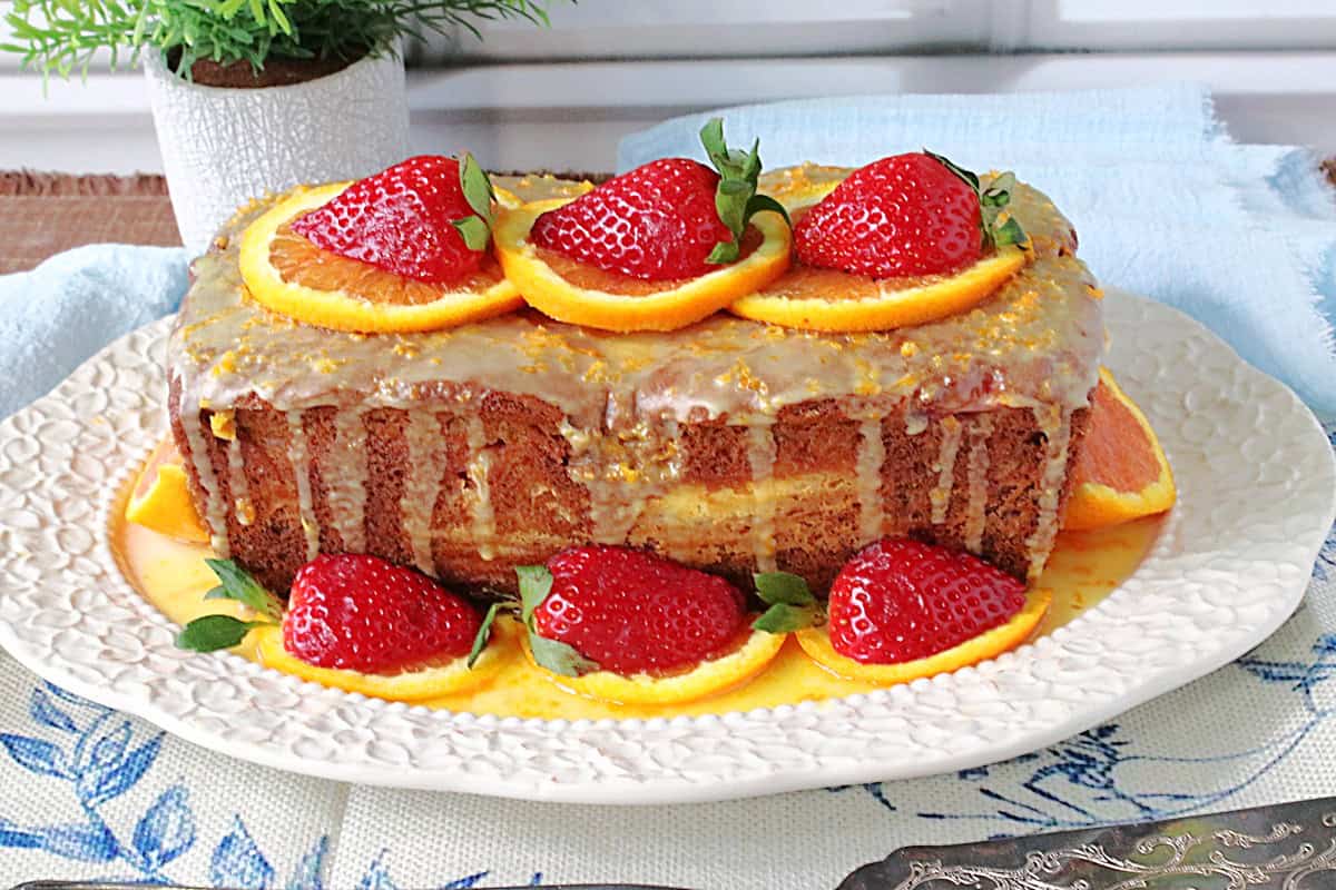 A blue and white table cloth with a white platter holding a glazed orange and strawberry loaf cake.