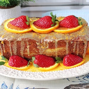 A beautiful orange and strawberry loaf cake that's been garnished with orange slices and strawberries on a pretty white platter.