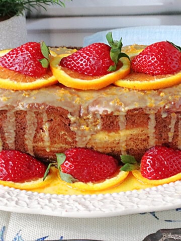 A beautiful orange and strawberry loaf cake that's been garnished with orange slices and strawberries on a pretty white platter.