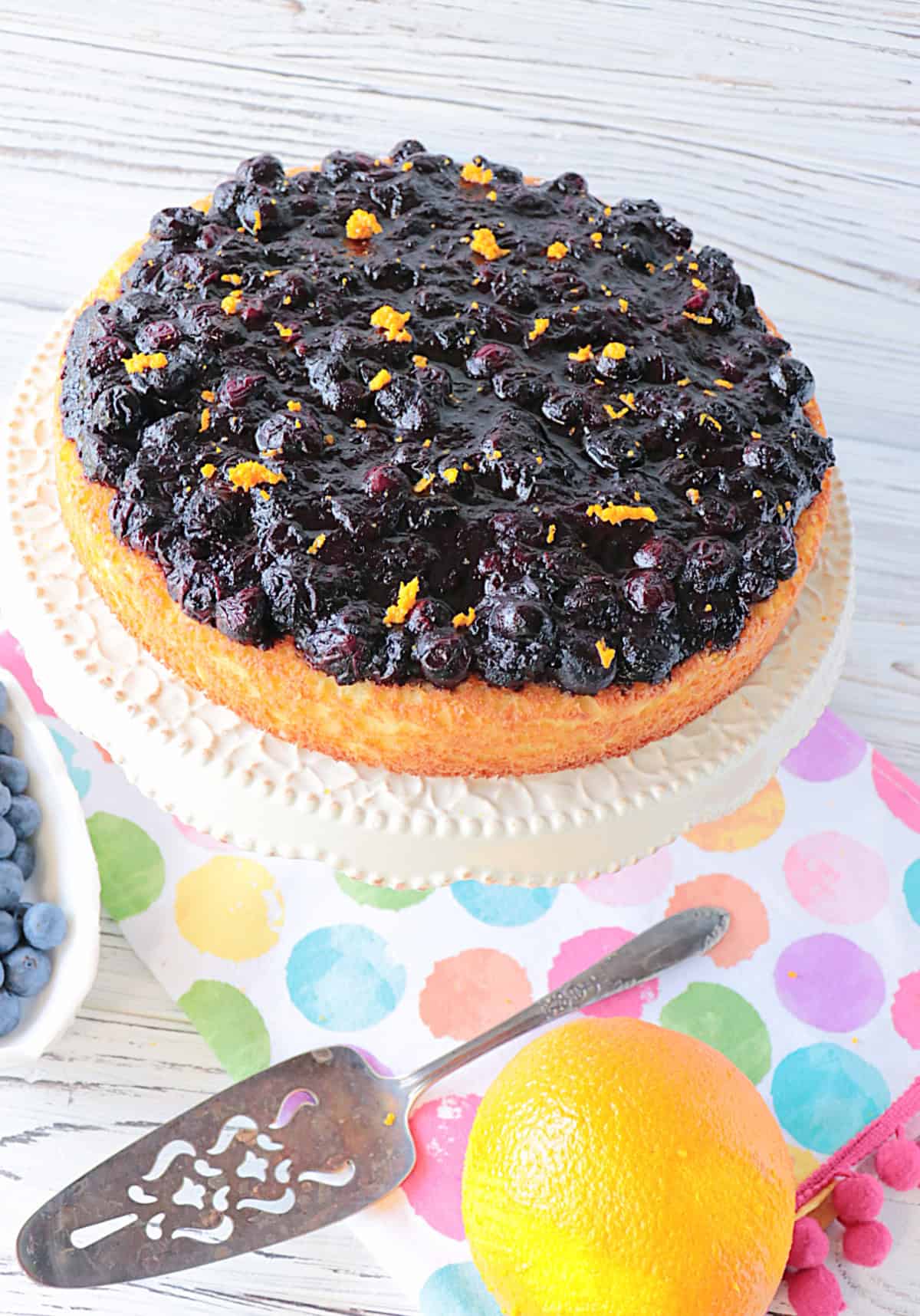 An overhead photo of a blueberry compote topped almond cake on a cake stand with a polka dot napkin.