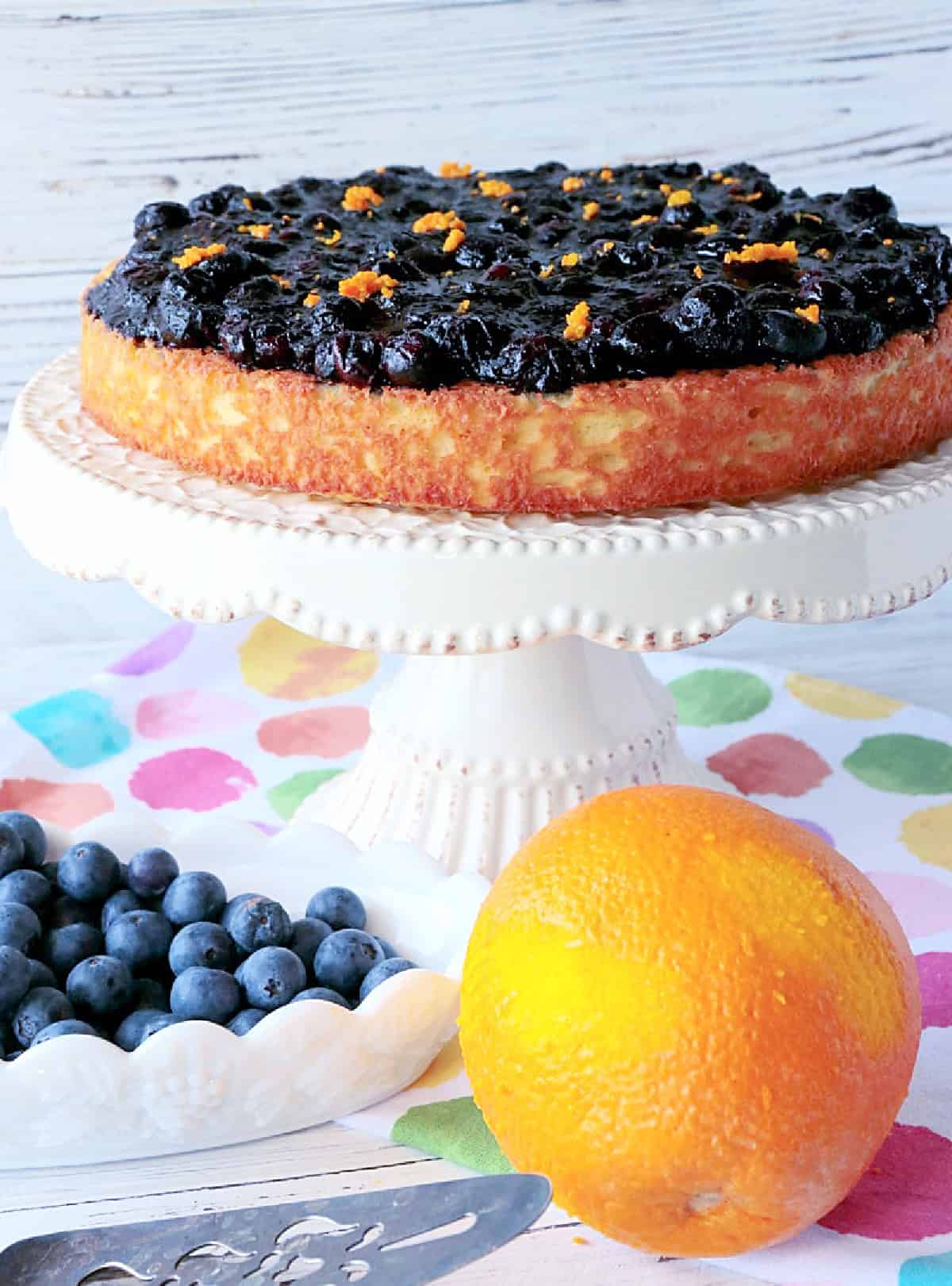 An almond cake topped with blueberry compote and orange zest on a pretty white cake stand.