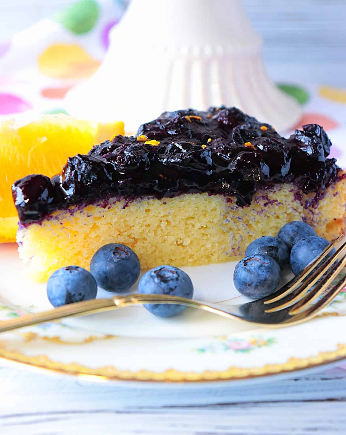 A moist looking slice of keto almond cake on a pretty plate with a fork and fresh blueberries.