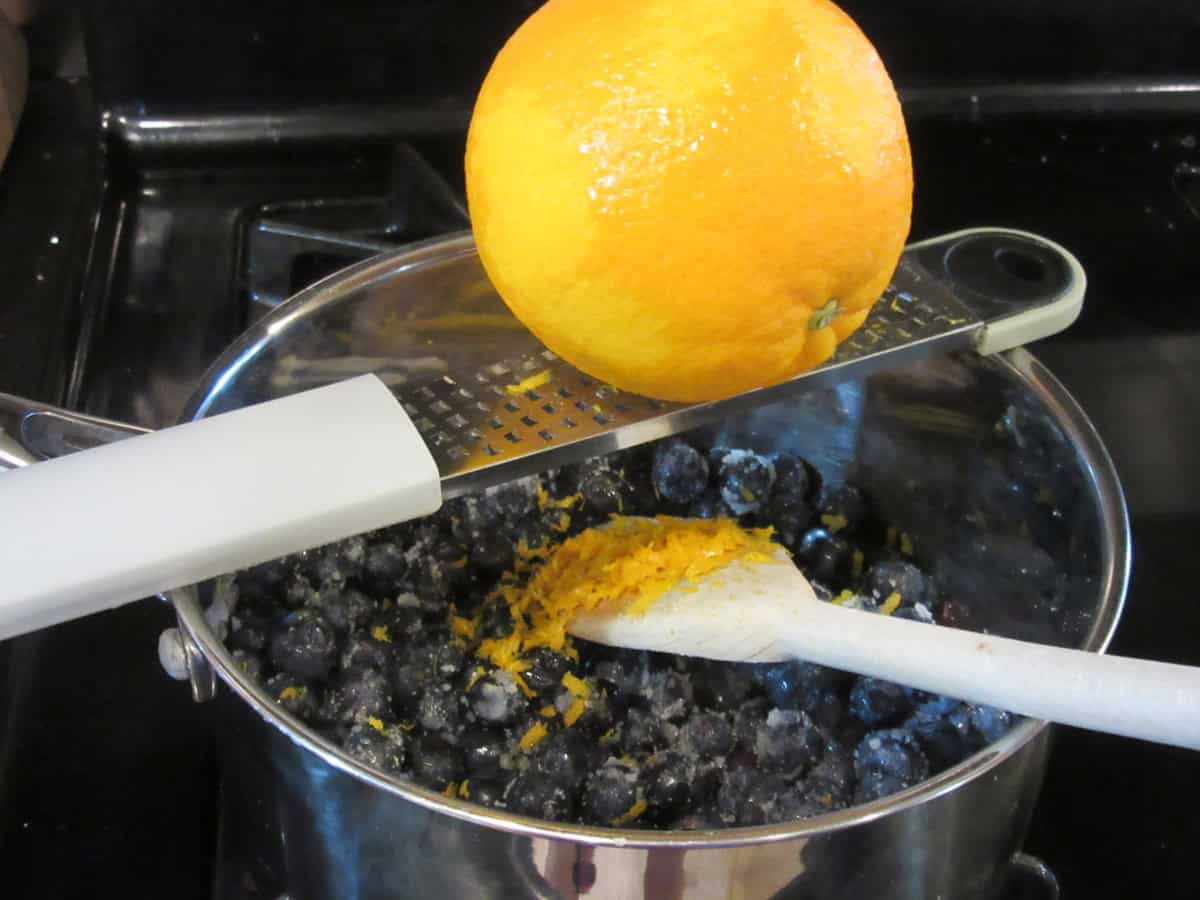 Blueberries in a saucepan with orange zest being added.