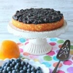 A very pretty almond cake topped with blueberry compote sitting on a white cake stand.