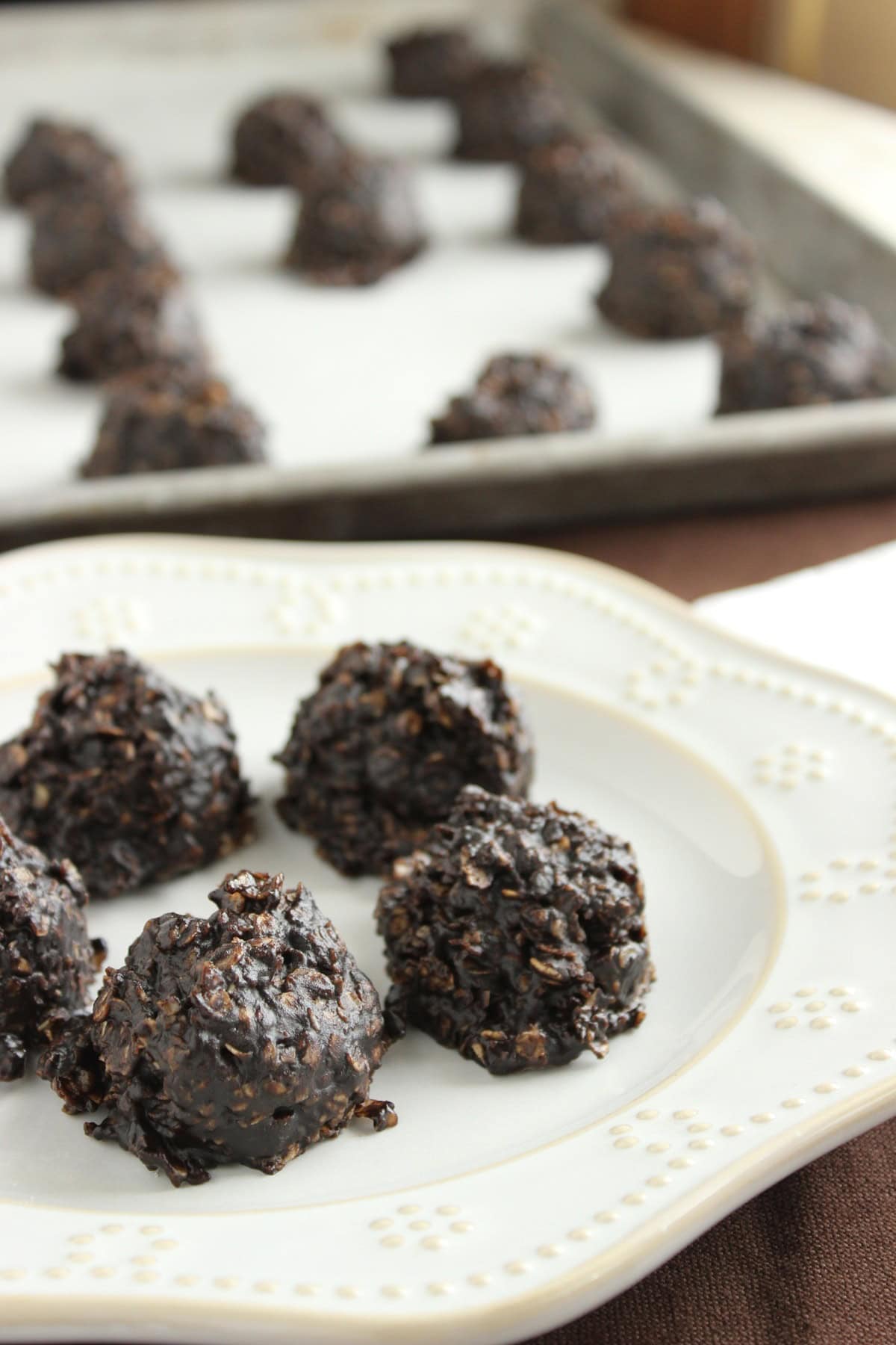 A baking sheet in the background and a white plate in the foreground all holding no bake chocolate and peanut butter drop cookies.