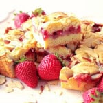 A bunch of cut strawberry cobbler bars stacked on a white plate surrounded by fresh strawberries.