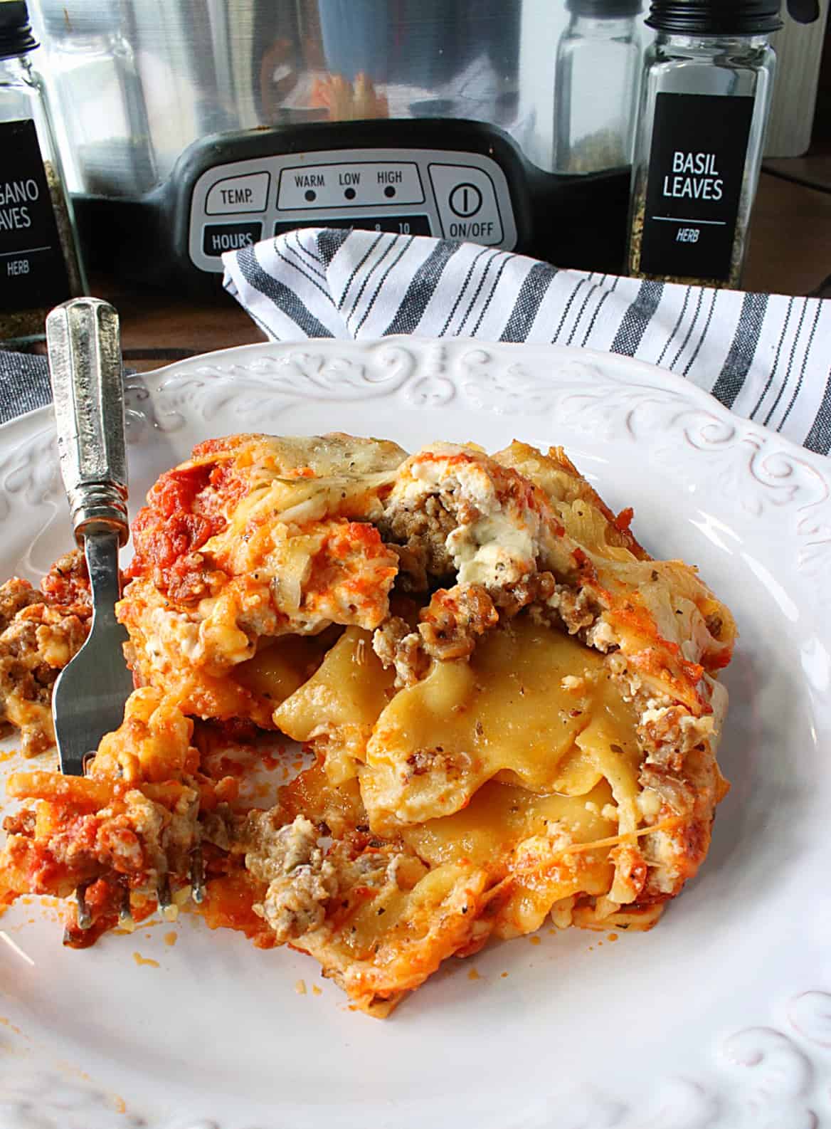 A slow cooker filled with lasagna in the background and a serving of lasagna on a white plate in the foreground.