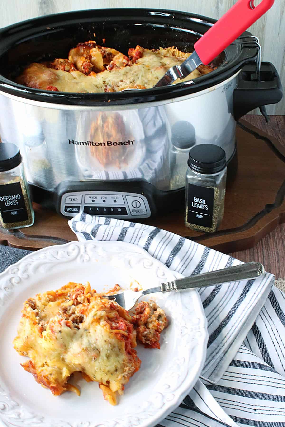 A silver slow cooker in the background and a serving of cheesy lasagna on a plate in the foreground.