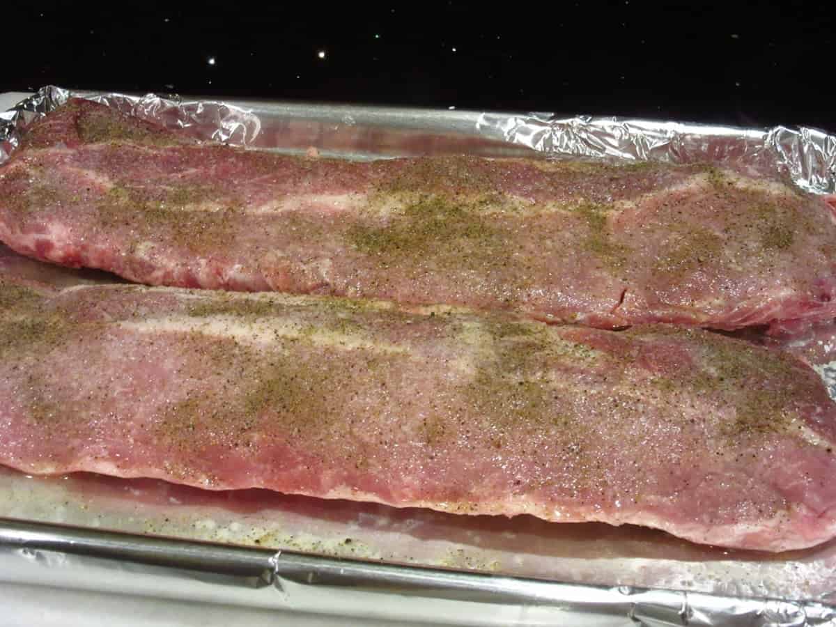 Two seasoned slabs of baby back ribs on an aluminum foil covered baking sheet.