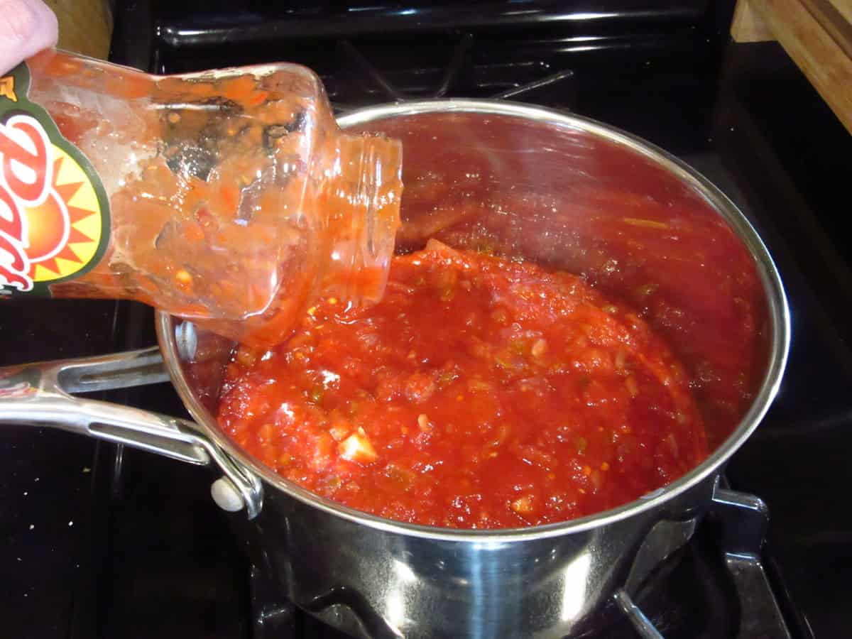 A jar of salsa being added to a saucepan on top of the stove.
