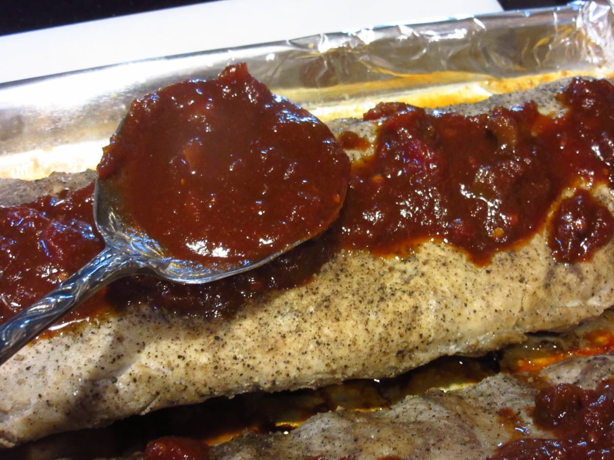Salsa BBQ sauce being spooned on the top of a partially cooked baby back rib.