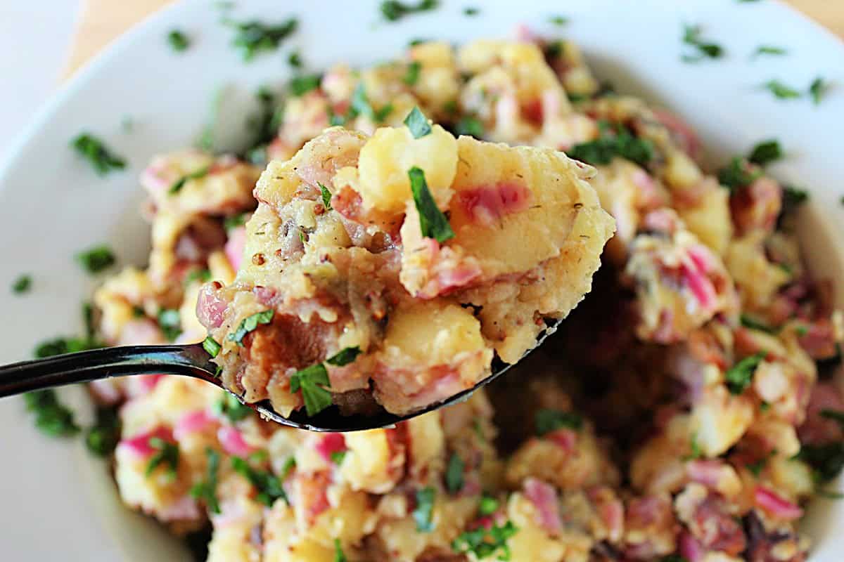 A spoonful of German potato salad being held over a bowl of the same salad.