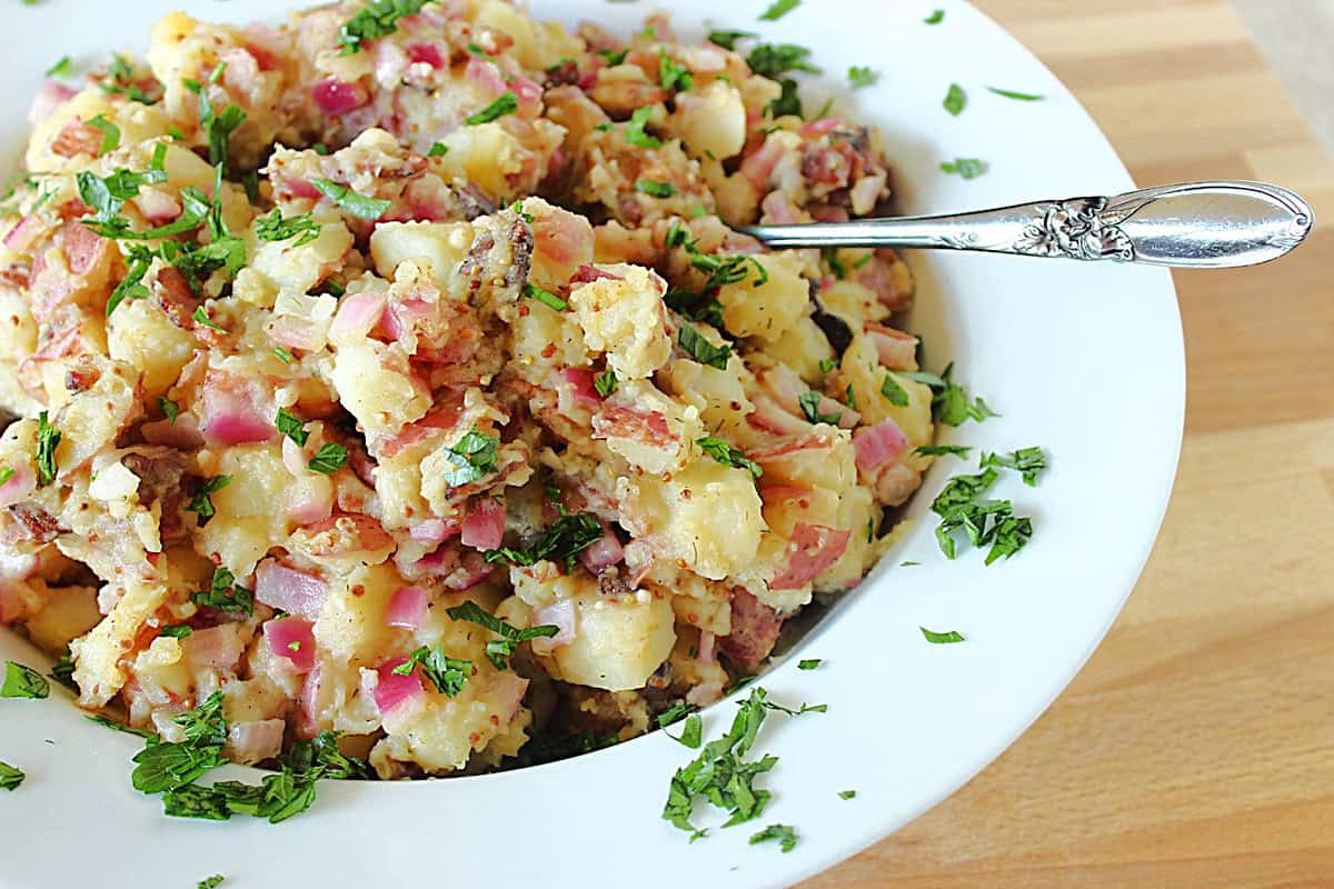 A silver serving spoon sticking out of a white bowl filled with traditional German Potato Salad.