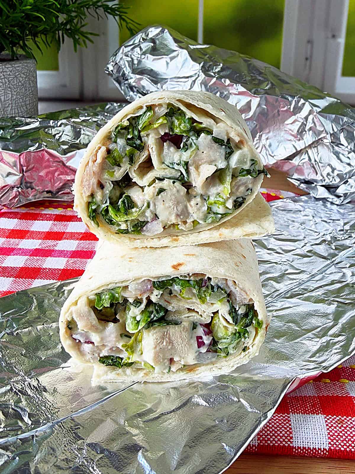 Foil wrapped chicken Caesar tortilla sandwiches on a red and white checked napkin.