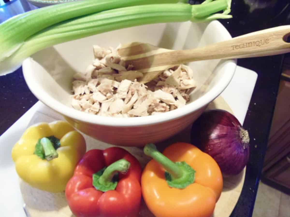 Colorful bell peppers and a red onion surrounding a bowl of diced rotisserie chicken along with a rib of celery on top of the bowl.