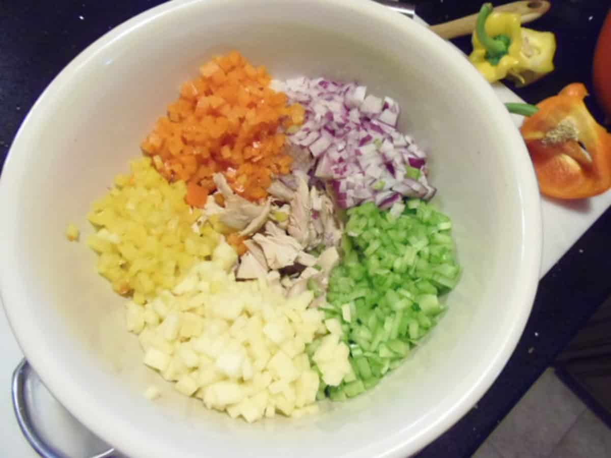 Finely diced, bell peppers, red onion, apple, and celery in a bowl of chopped, cooked chicken to make chicken salad.