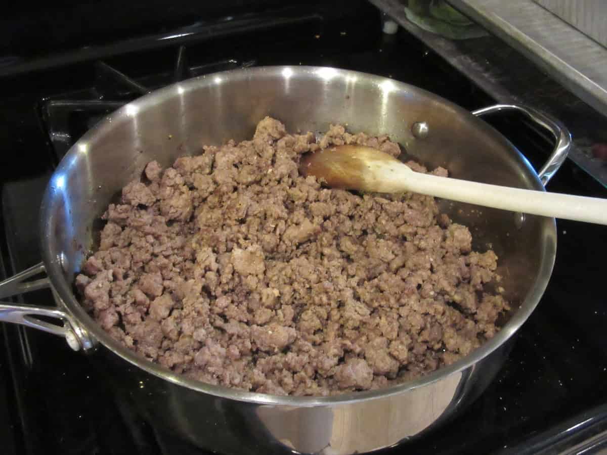 Two pounds of cooked and browned ground lamb in a large silver skillet on the stovetop along with a wooden spoon.