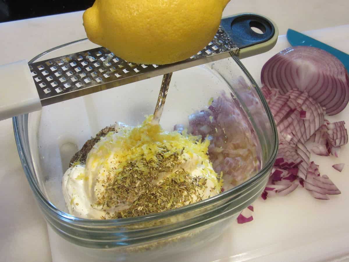 A whole lemon being zested over a bowl containing Greek tzatziki sauce.