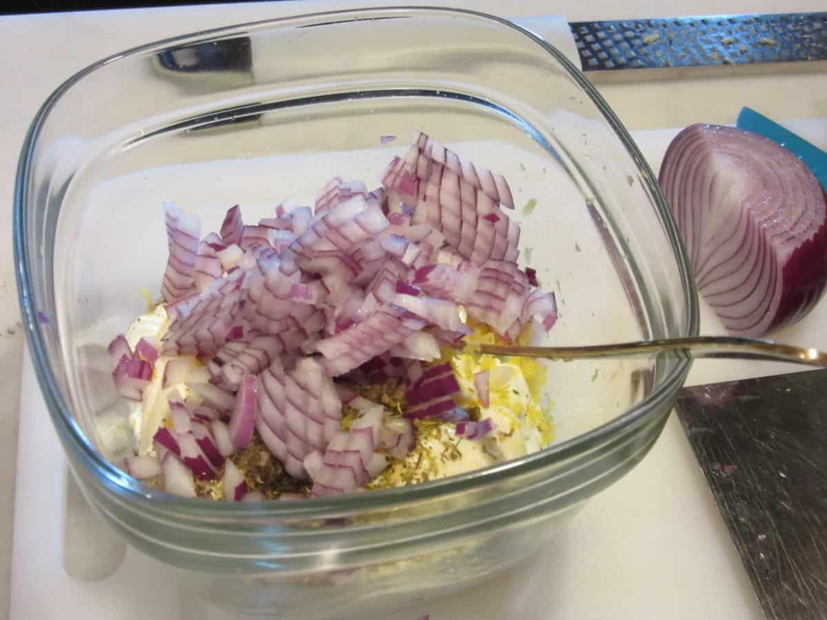 Diced red onion in a glass bowl with the makings of Greek tzatziki sauce along with a spoon.