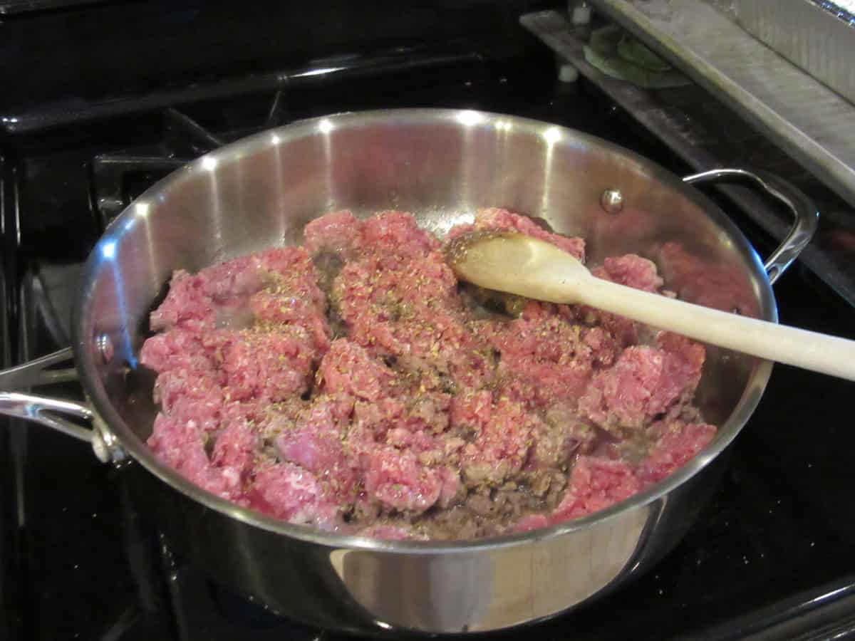 A large high-sided skillet on the stovetop browning two pounds of ground lamb along with seasonings.