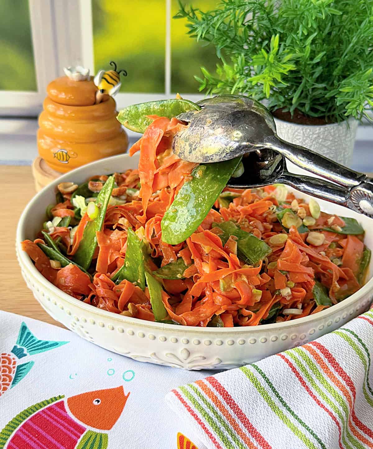 A pair of silver tongs serving up a shredded carrot and snow pea salad.