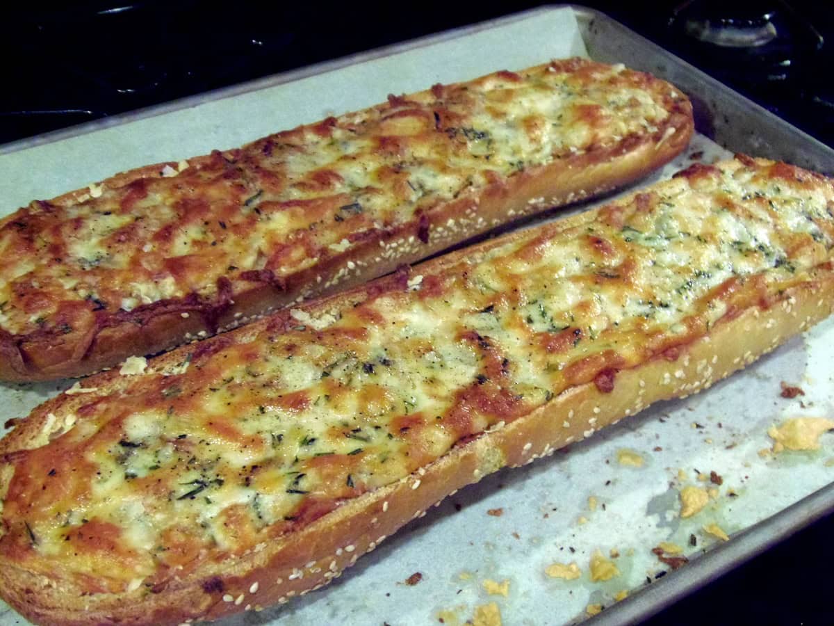 Two golden brown cheese topped garlic bread halves topped with rosemary on a baking sheet.