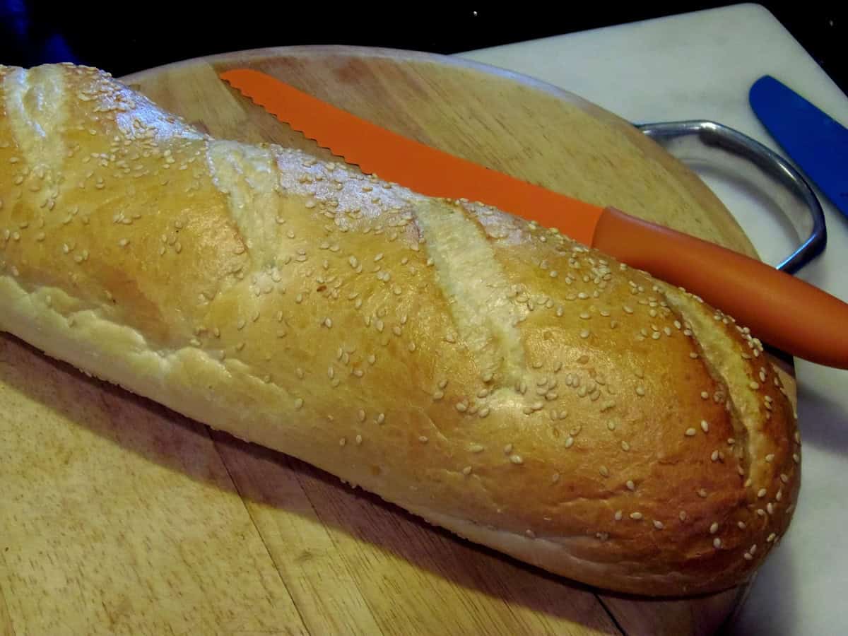 A whole loaf of seeded Italian bread on a wooden cutting board with an orange serrated knife on the side.