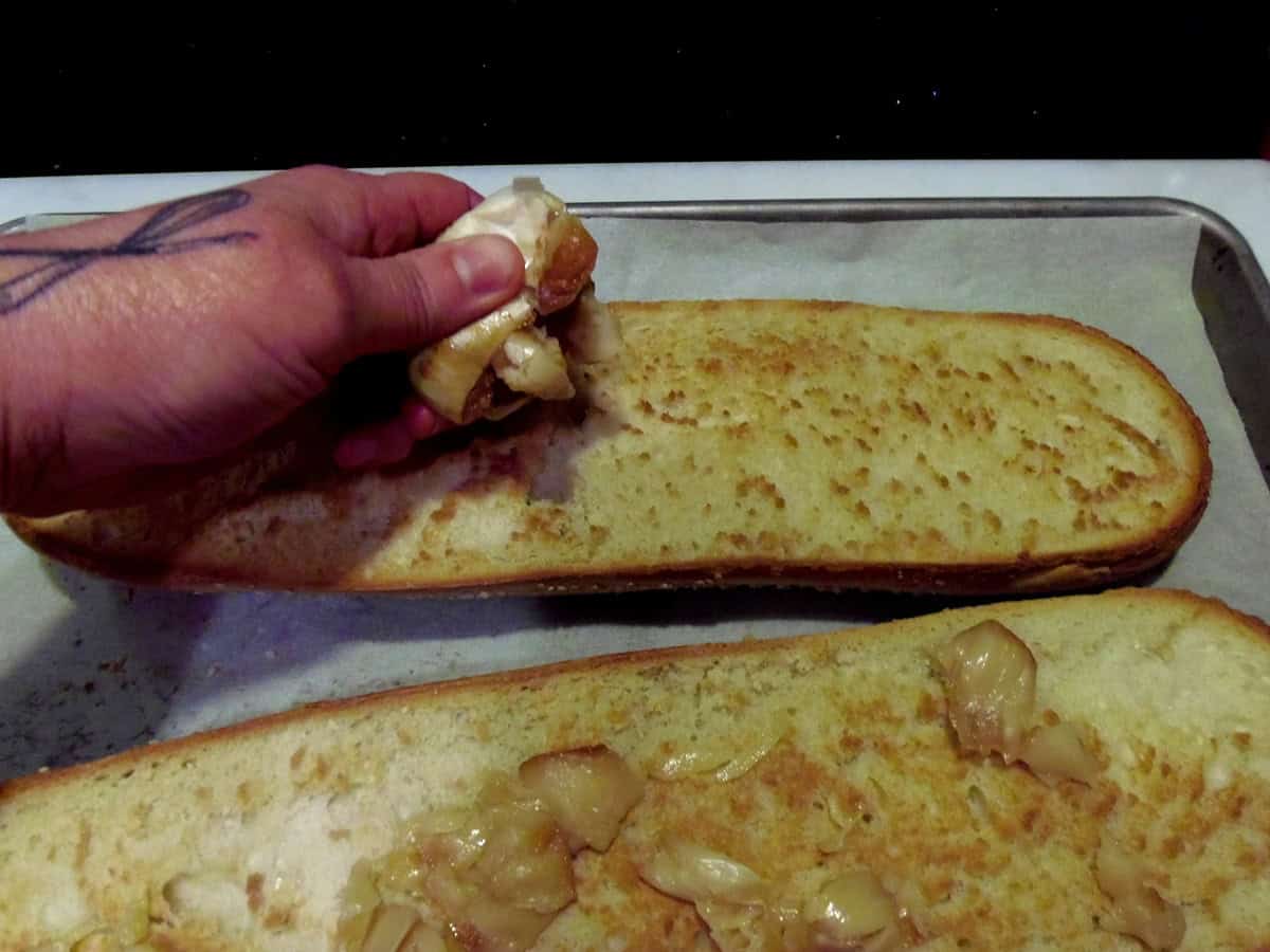A hand squeezing soft, roasted garlic out onto a toasted half of Italian bread.
