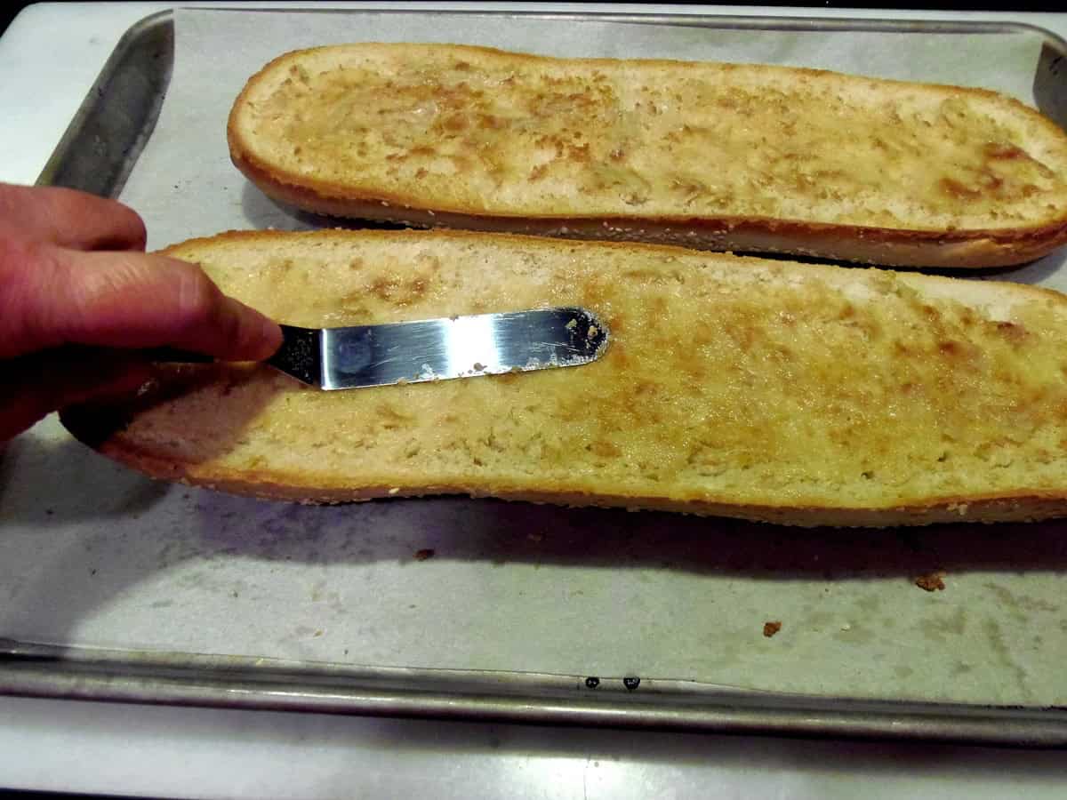 An offset spatula spreading roasted garlic bread on one half of a toasted Italian bread loaf.