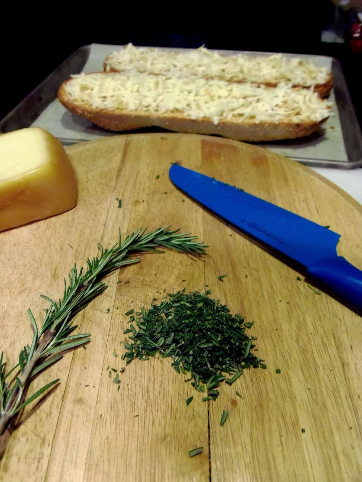 Finely minced fresh rosemary on a cutting board with a blue knife and some cheese topped bread in the background.