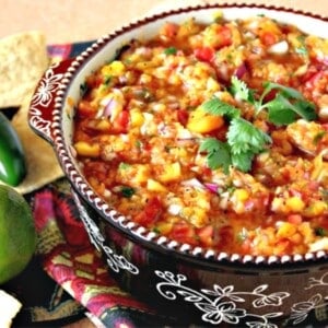 A colorful peach salsa with jalapenos and cucumbers topped with a cilantro sprig in a black bowl.