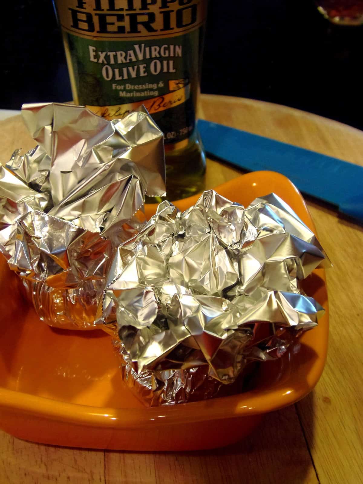 Two foil packets containing garlic bulbs in an orange dish with a bottle of olive oil in the background.