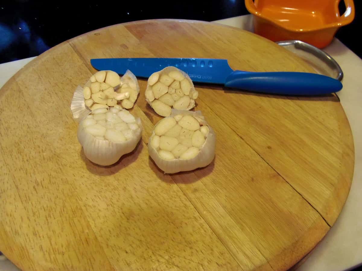 Two bulbs of garlic with the tops cut off on a wooden cutting board with a blue knife on the side.