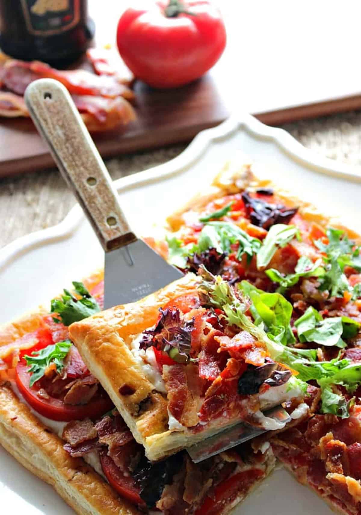 Whether you serve this delectable BLT Puff Pastry Tart with a Balsamic Drizzle for an appetizer or main course, you're going to love the fresh tomato flavor topped by oodles of crispy, smoked bacon. - Kudos Kitchen by Renee A serving spatula with a square of baked BLT puff pastry appetizer on top.