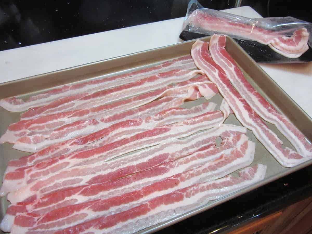 Strips of uncooked bacon laying on a baking sheet.
