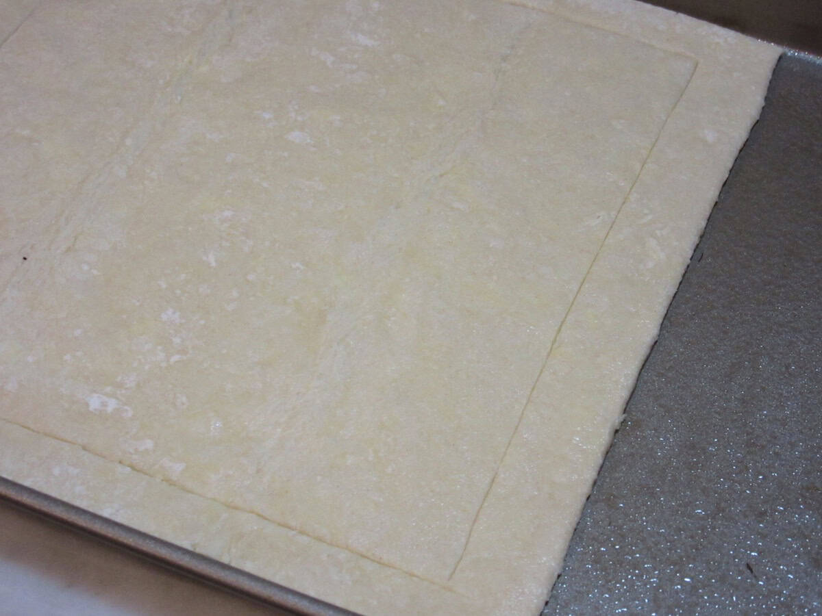 A scored frame border on a square of puff pastry dough before baking.