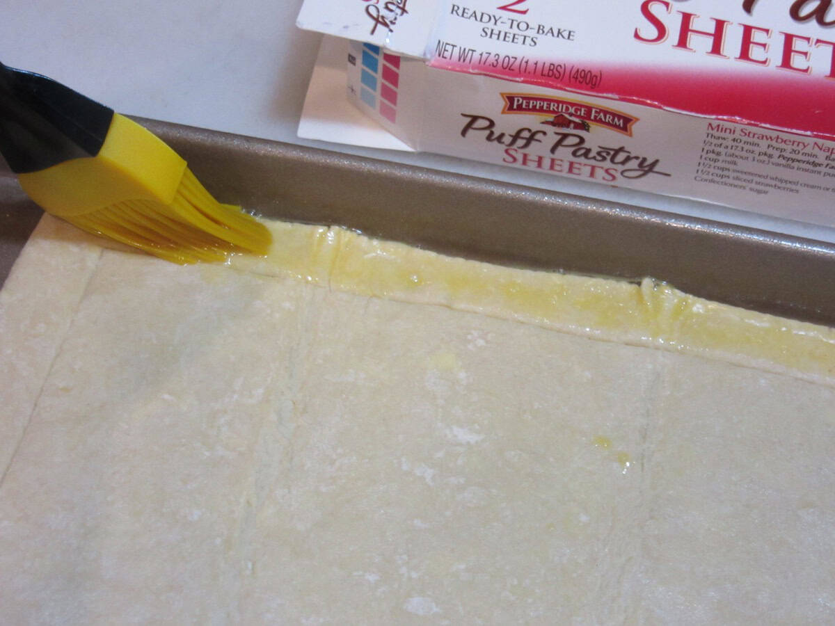 A pastry brush adding egg wash to the border of a sheet of puff pastry dough.