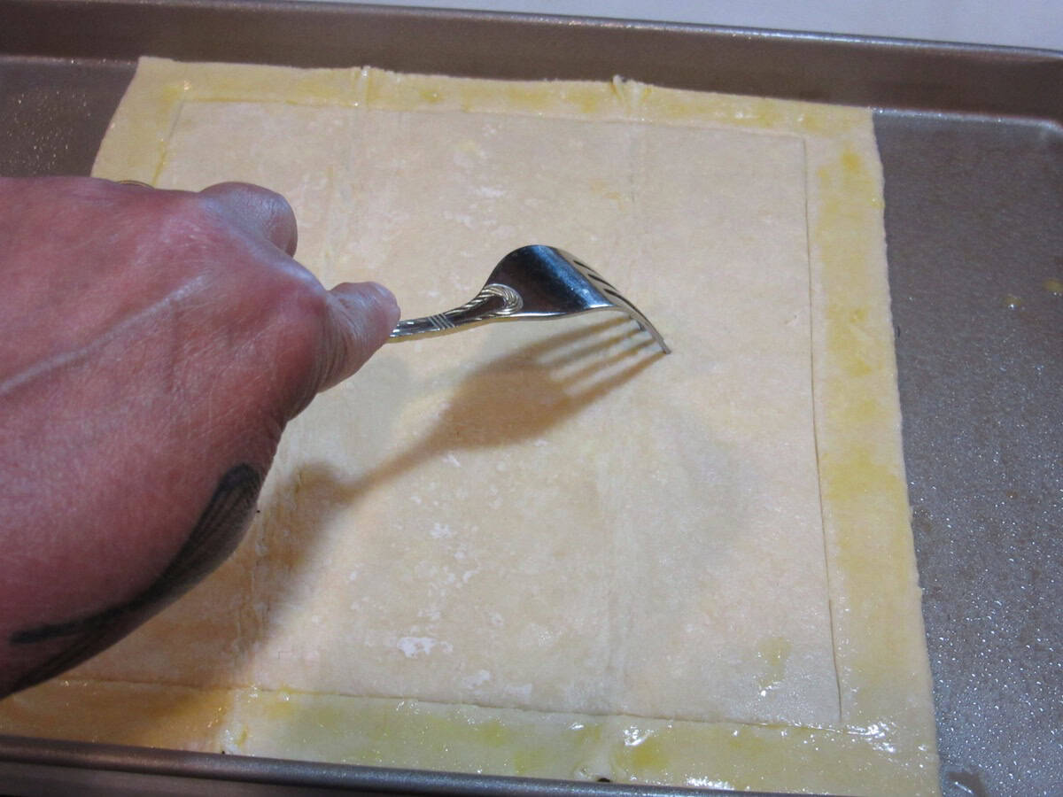 A hand holding a fork while poking holes into a sheet of puff pastry dough.